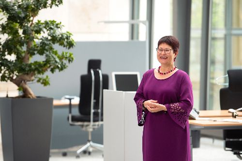 Frau Löffler steht lächelnd in einem Büro. Sie trägt ein lilanes Oberteil. Links von ihr steht ein kleiner Baum.