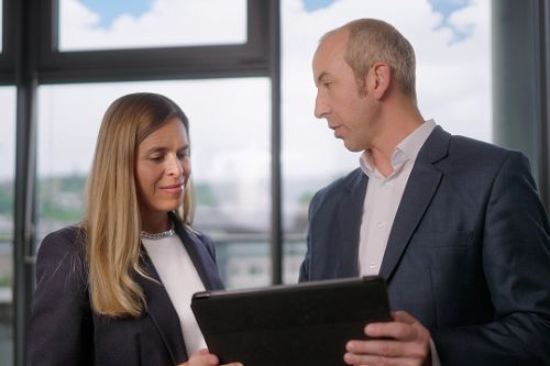 Zwei Personen betrachten gemeinsam ein Tablet in einem modernen Büro vor großen Fenstern mit Stadtblick.