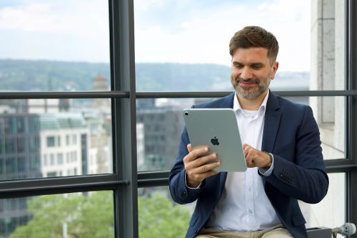 Eine Person sitzt mit einem Tablet in einem modernen Büro vor großen Fenstern.