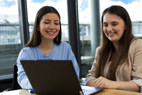 Zwei Personen arbeiten gemeinsam an einem Laptop in einem modernen Besprechungsraum und lächeln.
