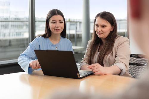 Zwei junge Frauen am Laptop in Besprechungsraum vor einer großen Fensterfront.
