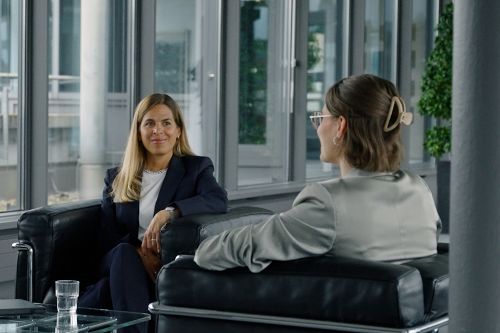 Zwei Personen sitzen sich in modernen schwarzen Ledersesseln in einem hellen Besprechungsraum mit großen Fenstern gegenüber.