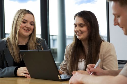 Zwei junge Frauen vor einem Laptop, ein junger Mann gegenüber.