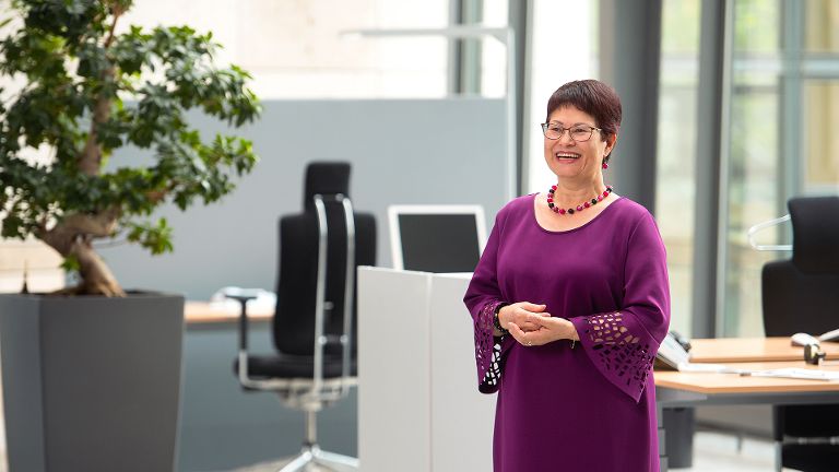 Frau Löffler steht lächelnd in einem Büro. Sie trägt ein lilanes Oberteil. Links von ihr steht ein kleiner Baum.