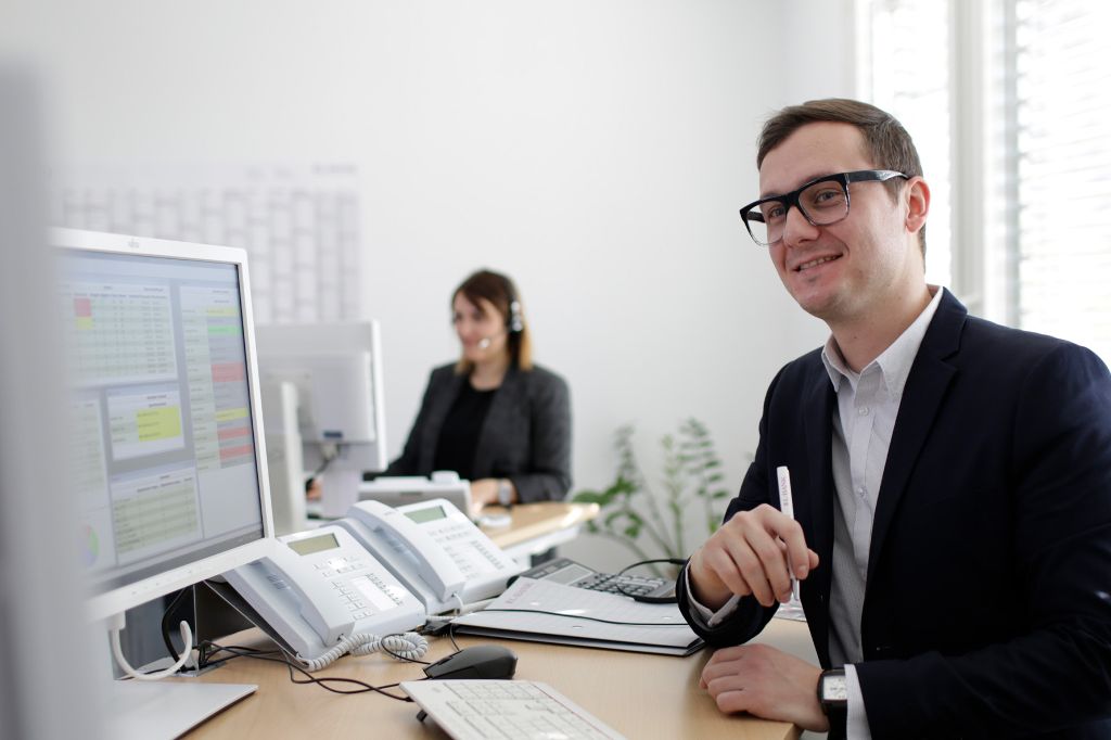 Ein Mann im Anzug steht mit einem Stift am Schreibtisch mit mehreren Computerbildschirmen.