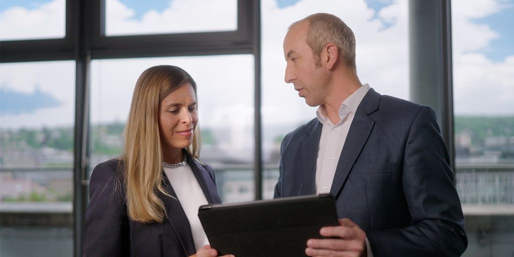 Zwei Personen betrachten gemeinsam ein Tablet in einem modernen Büro vor großen Fenstern mit Stadtblick.
