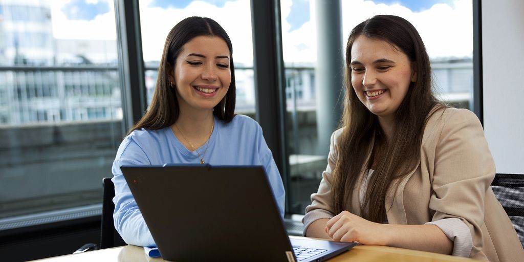Zwei Personen arbeiten gemeinsam an einem Laptop in einem modernen Besprechungsraum und lächeln.