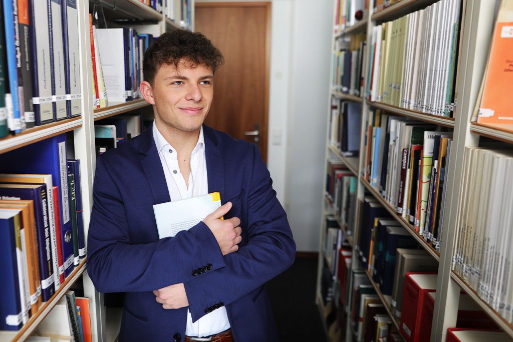 Eine Person steht zwischen Bücherregalen in einer Bibliothek und hält einen Ordner im Arm.