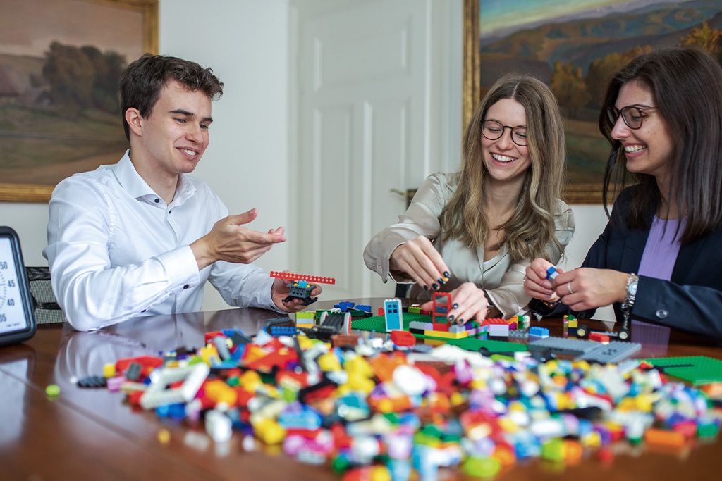 Drei Personen sitzen an einem Tisch und bauen gemeinsam mit bunten Lego-Bausteinen verschiedene Modelle.