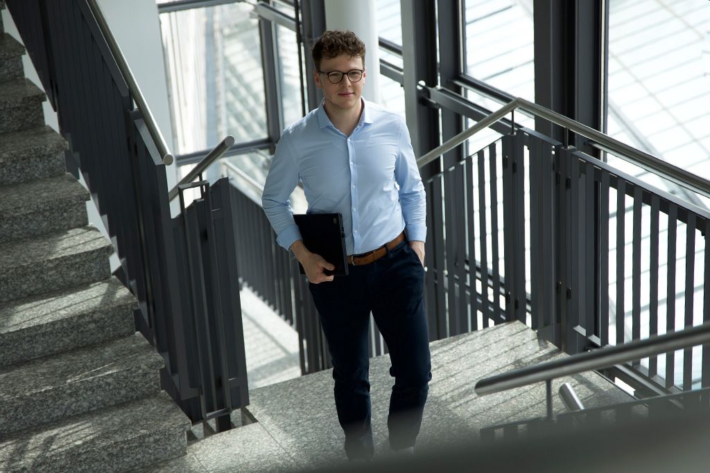 Eine junger Mann geht mit einem Tablet die Treppe in einem modernen Bürogebäude mit großen Fensterfronten hinauf.