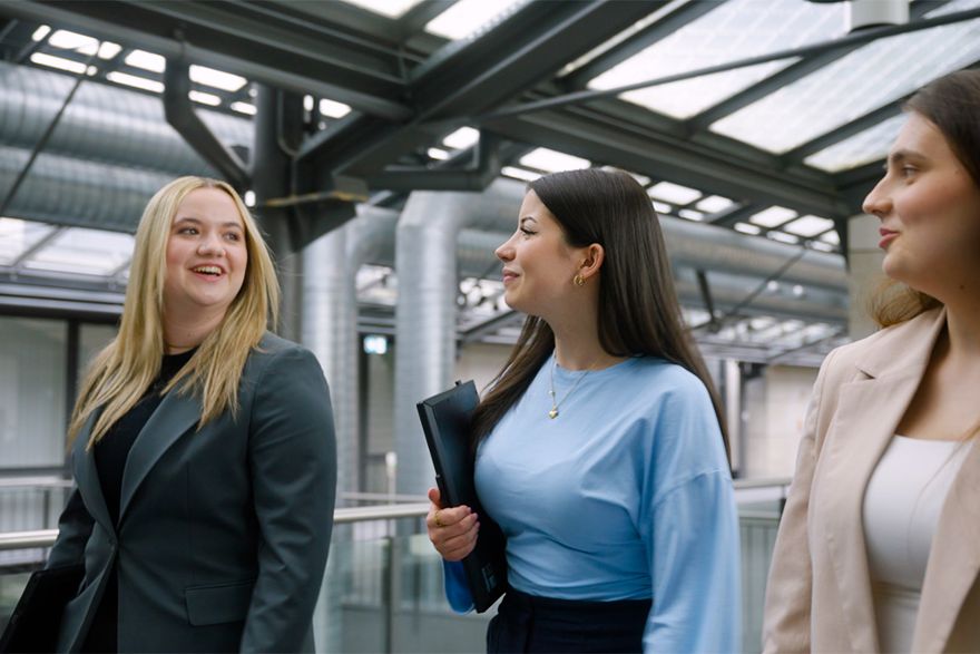 Drei junge Frauen gehen gemeinsam durch das Atrium eines modernen Bürogebäudes und unterhalten sich.