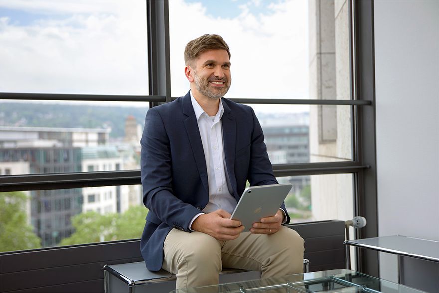 Mann in Anzug sitzt vor einem Fenster in einem Bürogebäude mit Tablet in der Hand und lächelt.