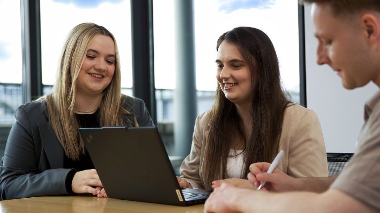 Zwei junge Frauen vor einem Laptop, ein junger Mann gegenüber.
