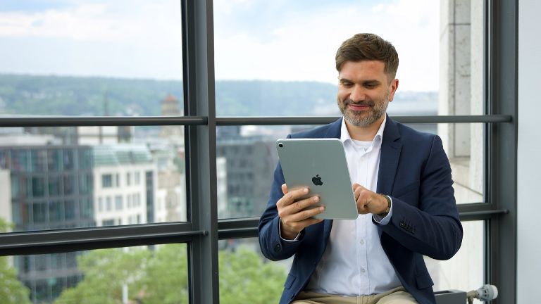 Eine Person sitzt mit einem Tablet in einem modernen Büro vor großen Fenstern.
