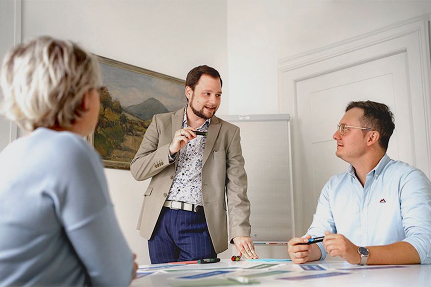 Drei Personen besprechen Unterlagen an einem Tisch, während eine Person stehend präsentiert.