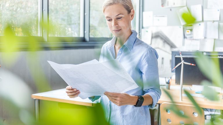 Eine lächelnde Frau liest ein Dokument in einem Büro, hinter ihr auf dem Schreibtisch steht ein Miniatur-Windrad