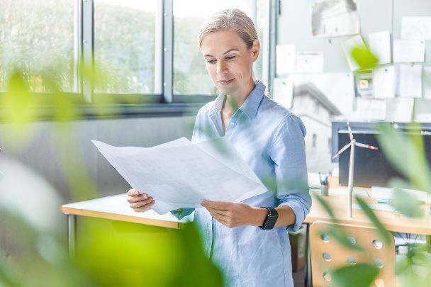 Eine lächelnde Frau liest ein Dokument in einem Büro, hinter ihr auf dem Schreibtisch steht ein Miniatur-Windrad