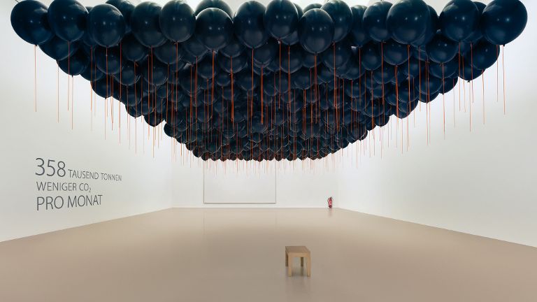 Mehrere hundert schwarze Luftballons schweben in einem kahlen weißen Raum. An der Wand steht 358000 Tonnen weniger CO2 pro Monat