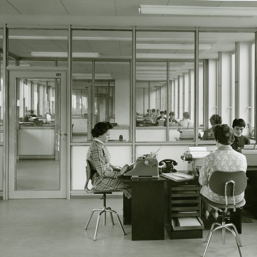 Büroräume im Stil der 1950er-Jahre, im Vordergrund sitzen drei Frauen jeweils an einer Schreibmaschine