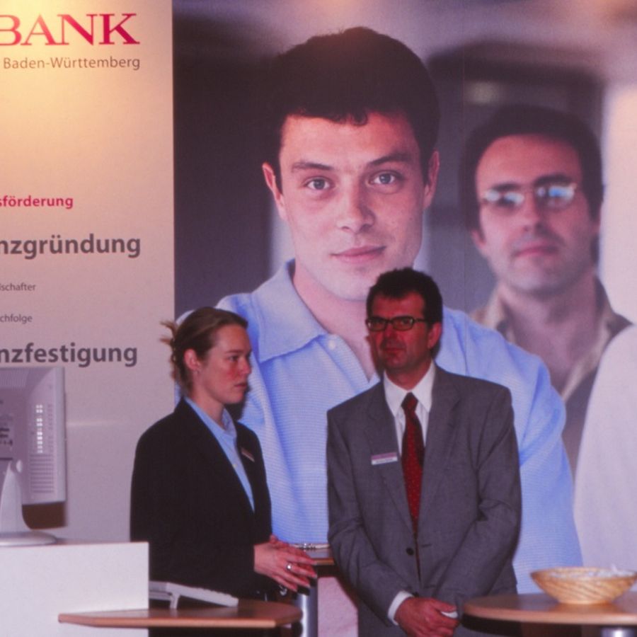 Eine Frau und ein Mann vor einem Aufsteller der L-Bank, auf dem mehrere Personen und die Worte Wirtschaftsförderung, Existenzgründung und Existenzfestigung zu sehen sind.