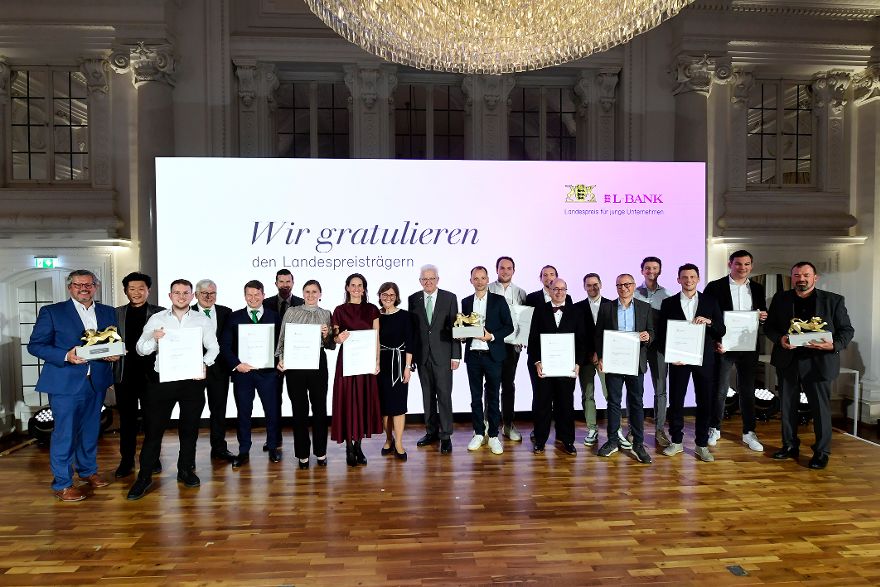Mehrere formell gekleidete Menswchen vor einem Aufstelller mit der Aufschrift Wir gratulieren den Landespreisträgern.